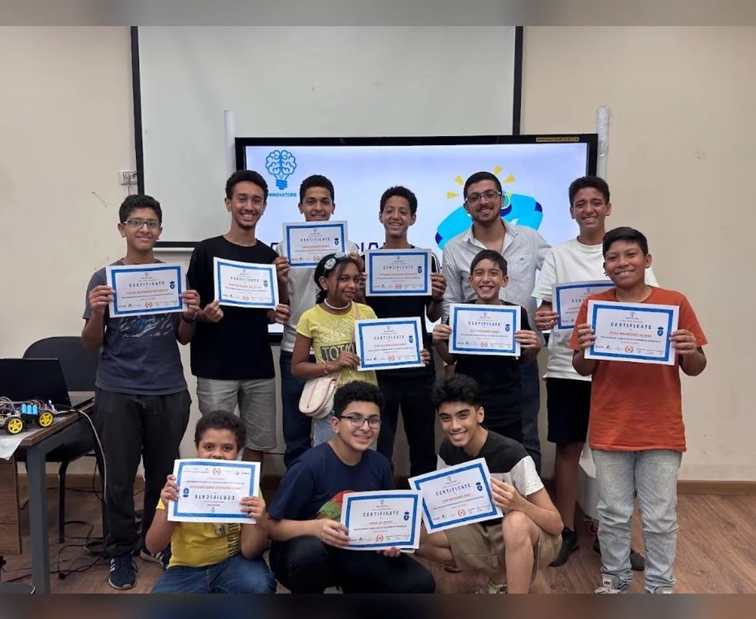Innovators Academy students holding their diploma certificates in the robotics lab, with a student-built robot car on the desk